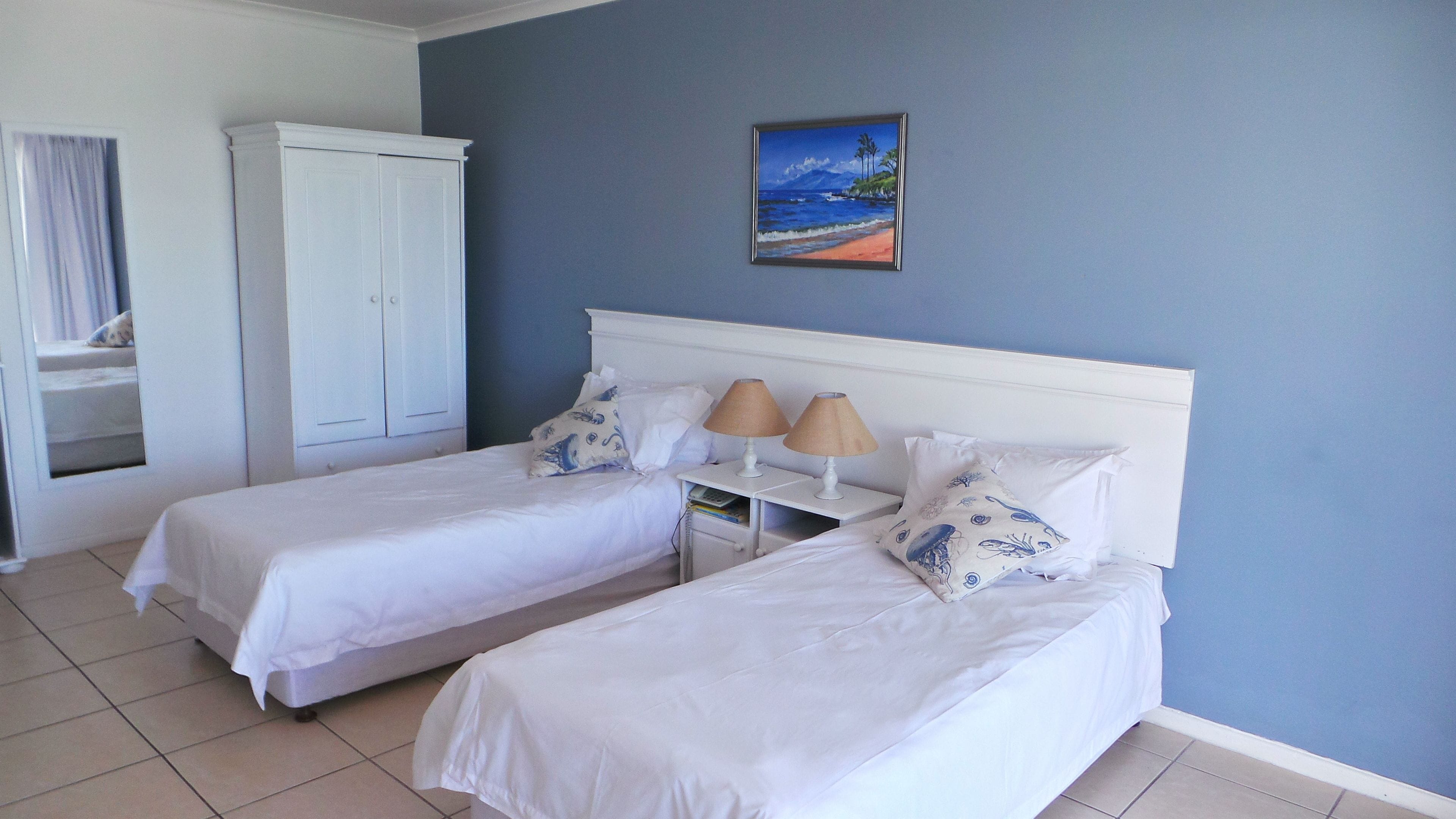 A hotel room with two single beds, a white wardrobe, a small bedside table with lamps, and a painting of a beach scene.