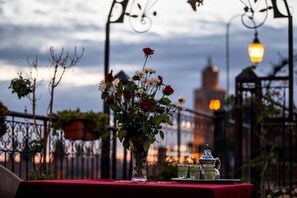 Une table recouverte d’une nappe rouge, un vase de fleurs et deux théières sur un balcon offrant une vue sur la ville.