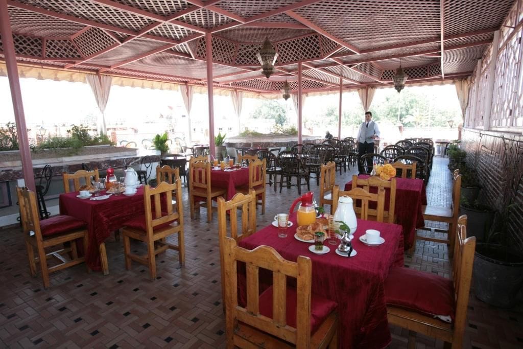 Un área de comedor al aire libre con mesas y sillas, cubierta por un toldo rojo, y una persona de pie al fondo.