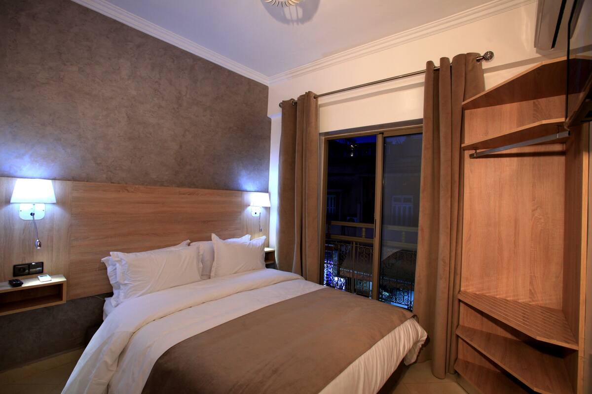 A hotel room with a large bed, two bedside lamps, a wooden headboard, a window with curtains, and a wooden wardrobe.
