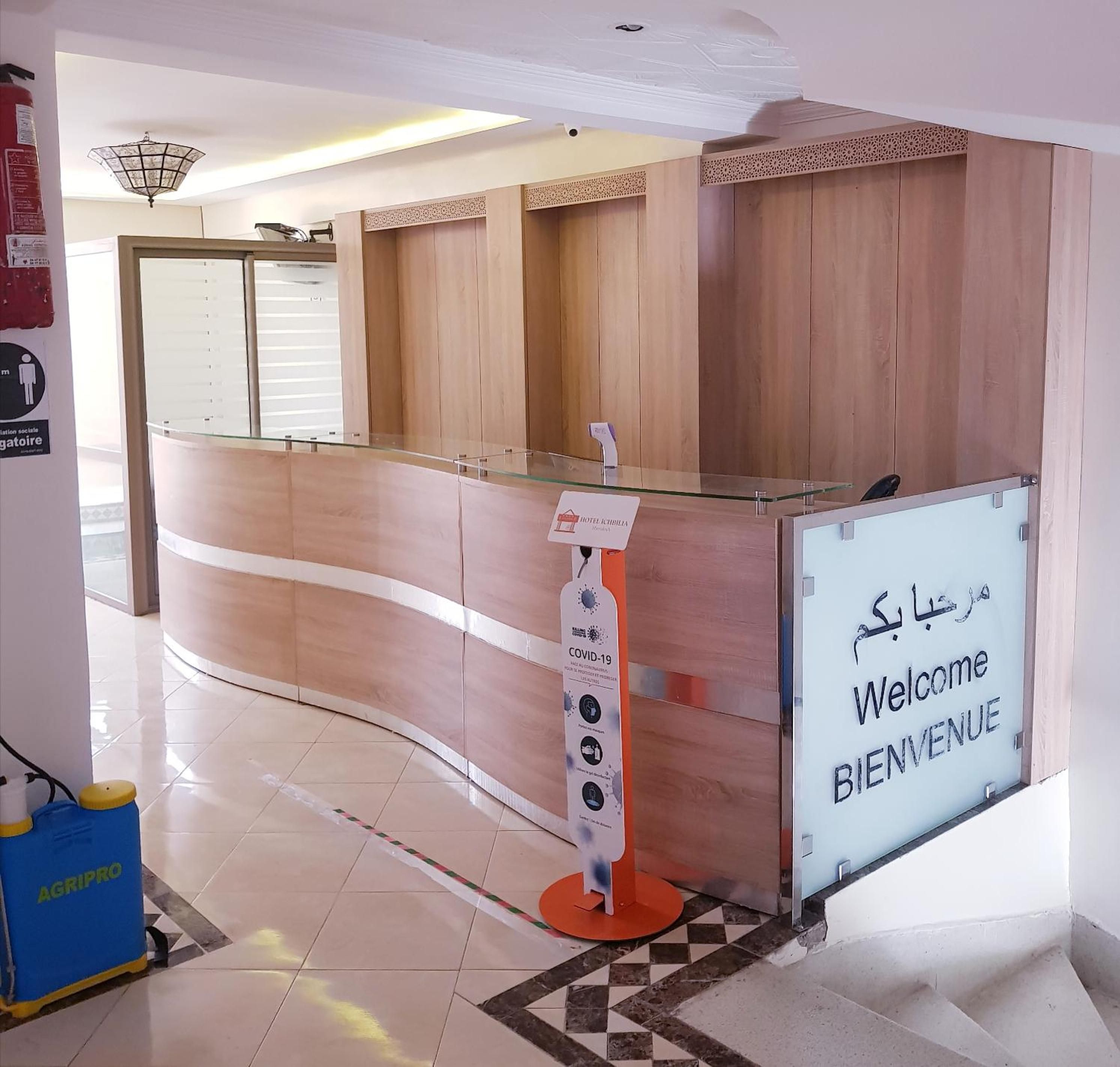 Balcão de recepção de hotel com divisória de vidro, placa de boas-vindas e suporte com informações sobre COVID-19.