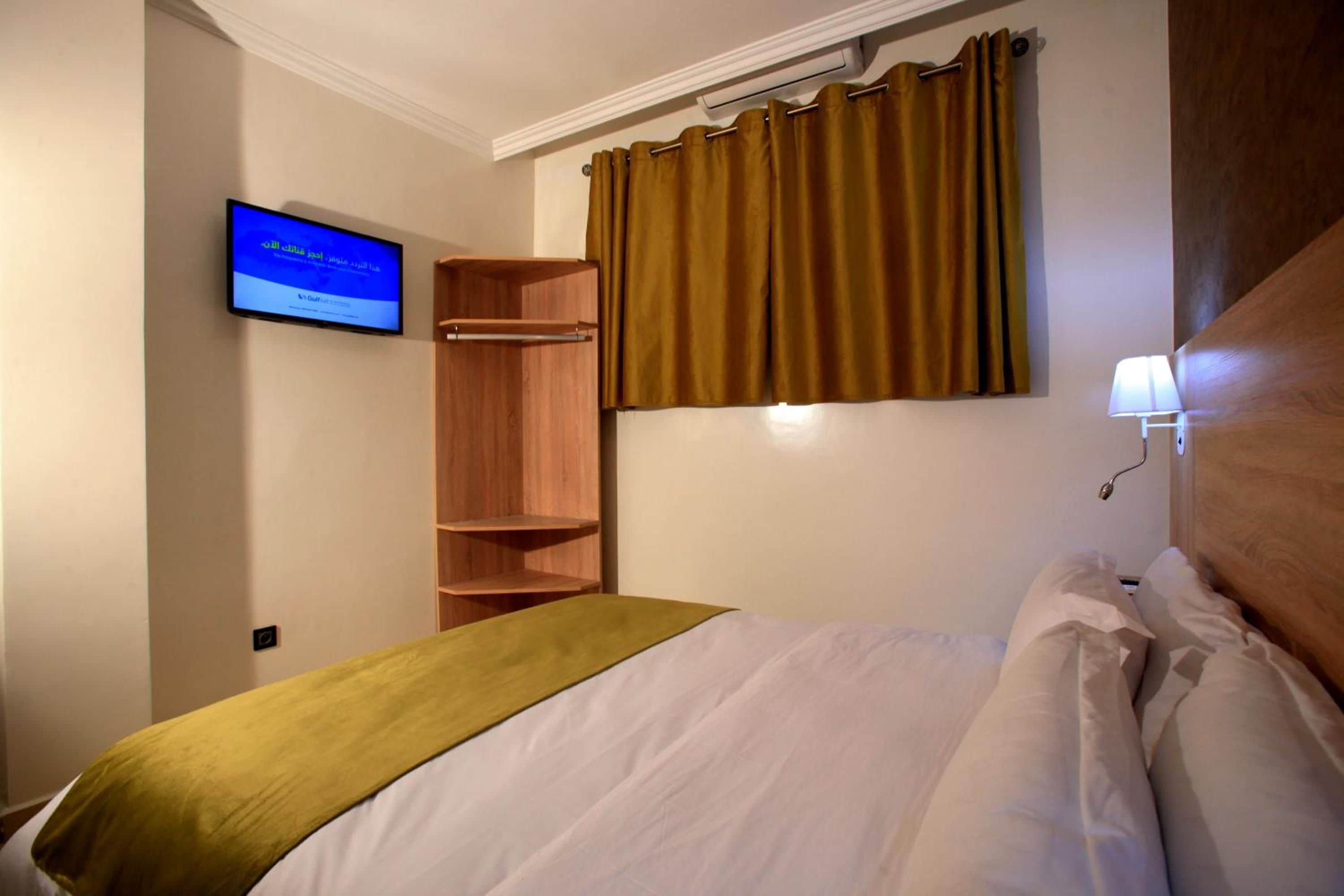 Una habitación de hotel con cama, televisor montado en la pared, armario de madera y ventana con cortinas.