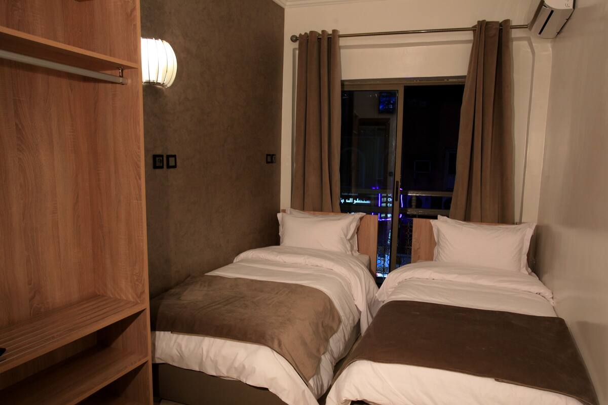 A hotel room with two beds, a wooden wardrobe, a window with curtains, and a view of a cityscape at night.
