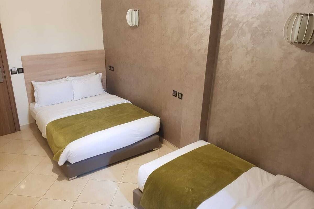 A hotel room with two beds, each with white bedding and a green throw. The room has a wooden headboard and beige walls.