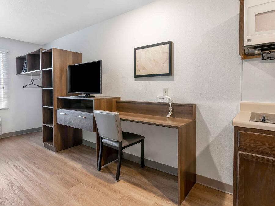 A compact hotel room with a wooden desk, a chair, a television, a framed picture, a built-in microwave, and a small bathroom area.
