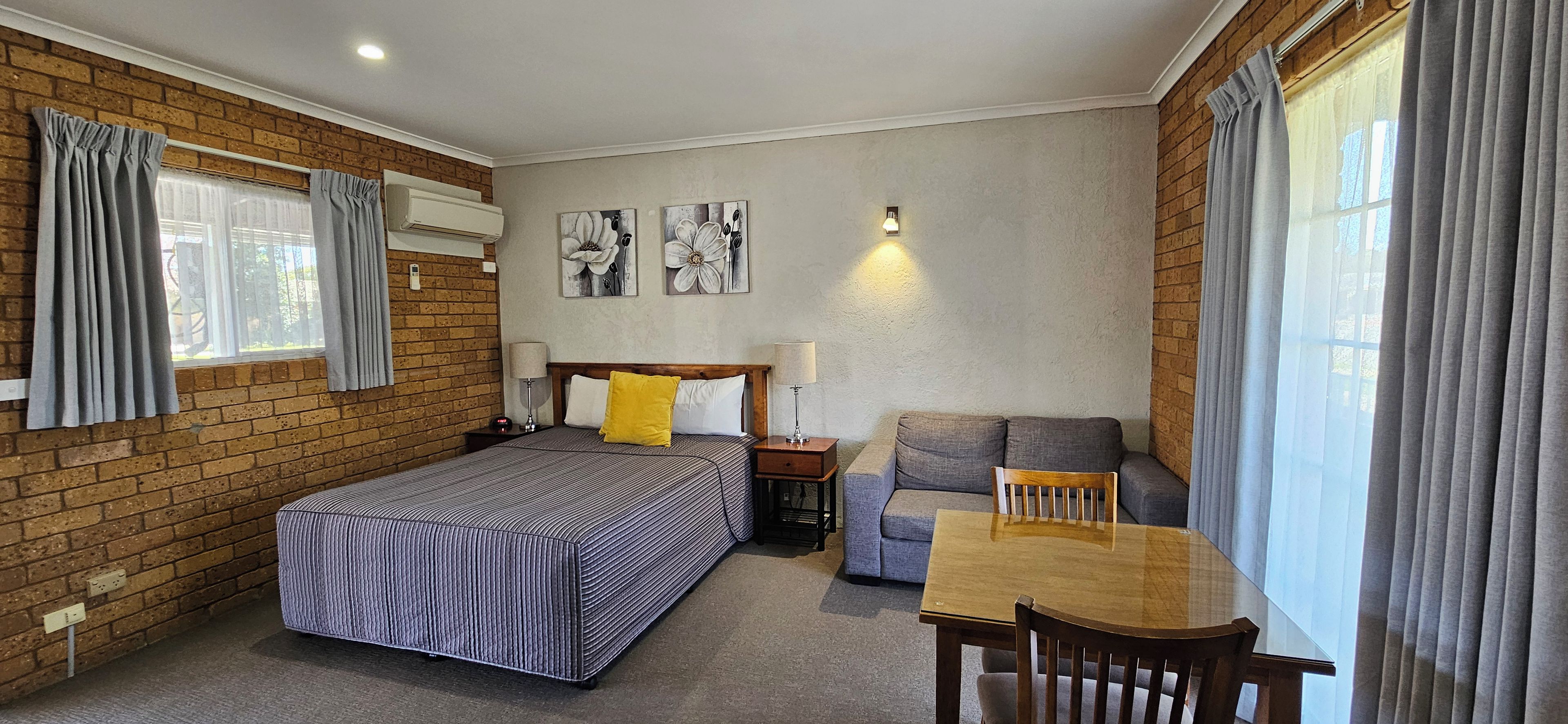 Une chambre d’hôtel avec un lit, un canapé, une table et une chaise. Il y a une fenêtre avec des rideaux, un climatiseur fixé au mur et deux lampes fixées au mur.