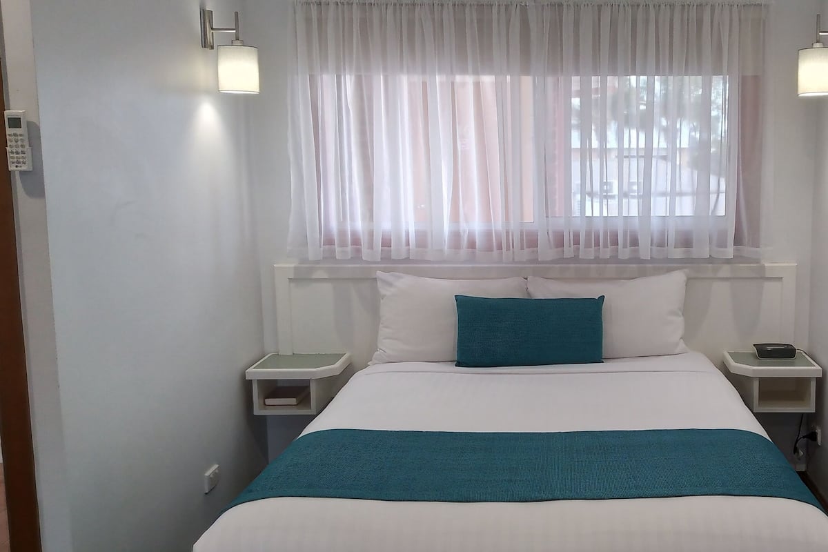A hotel room with a bed, white curtains, two bedside tables with lamps, and a window with sheer curtains.
