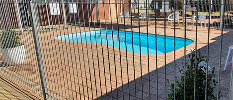A swimming pool surrounded by a metal fence, with a brick patio and white plastic chairs.