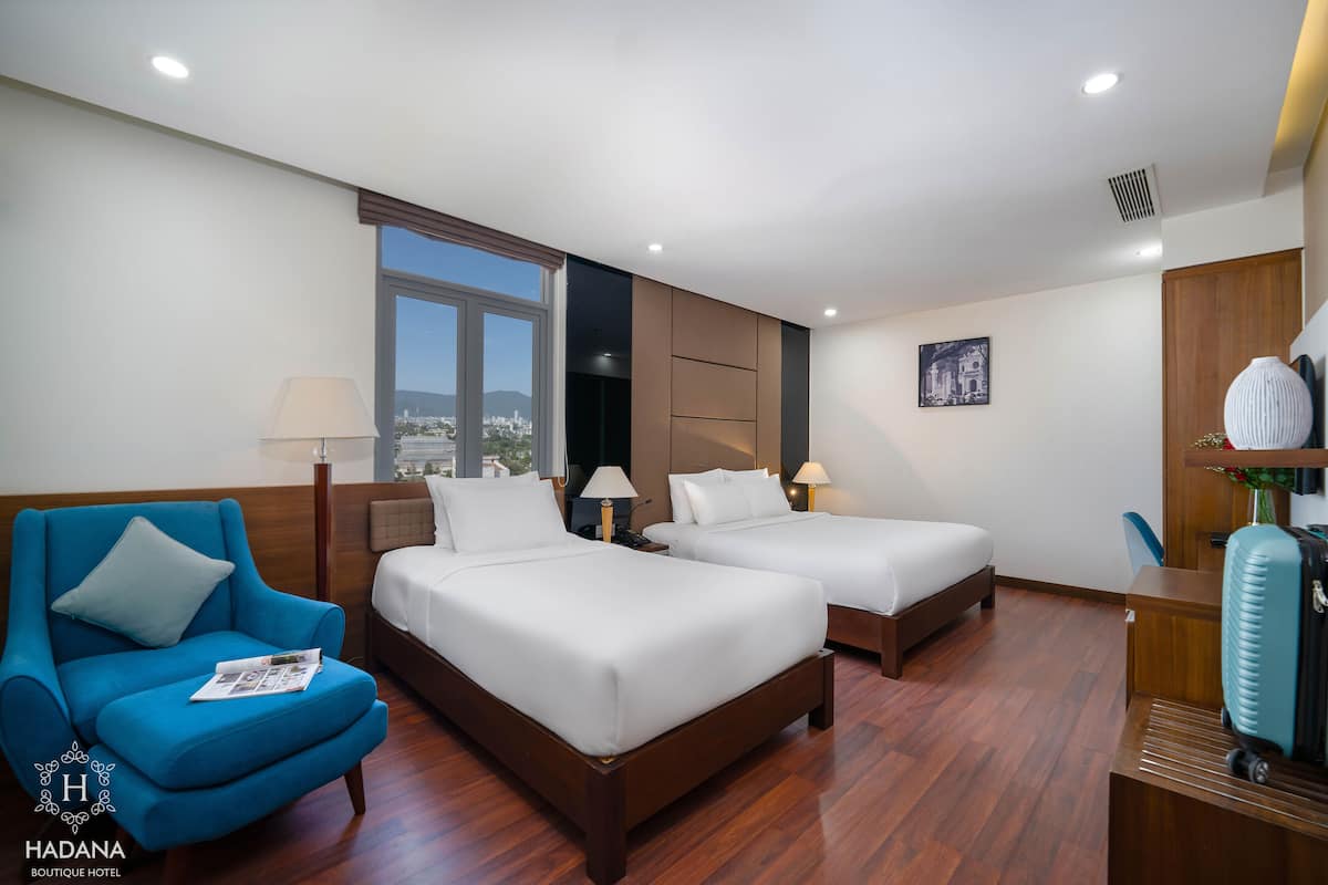 A hotel room with two beds, a blue armchair, a desk, and a view of the city.