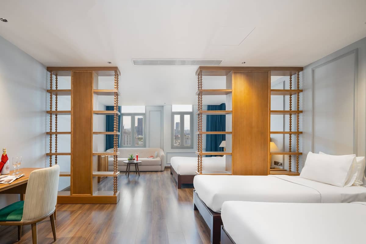 A hotel room with a wooden partition, a sofa, a bed, a desk with a chair, and a view of the city through large windows.
