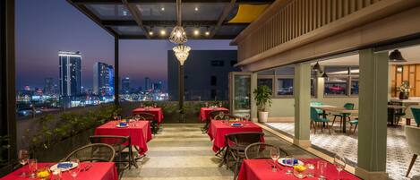 A rooftop dining area with tables set for dinner, overlooking a city skyline at dusk.