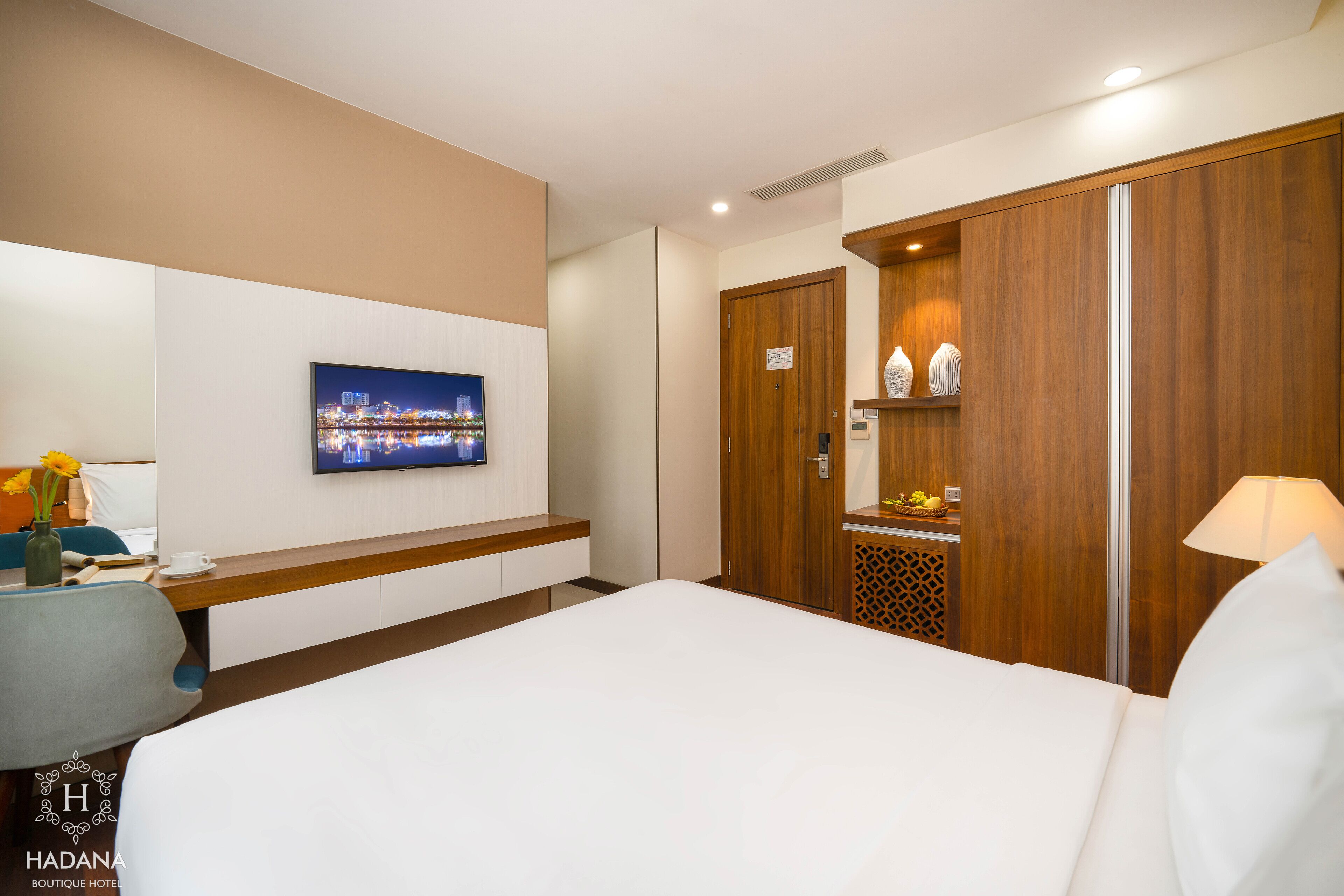Una habitación de hotel con cama, televisión montada en la pared, armario de madera y una mesita de noche con lámpara.