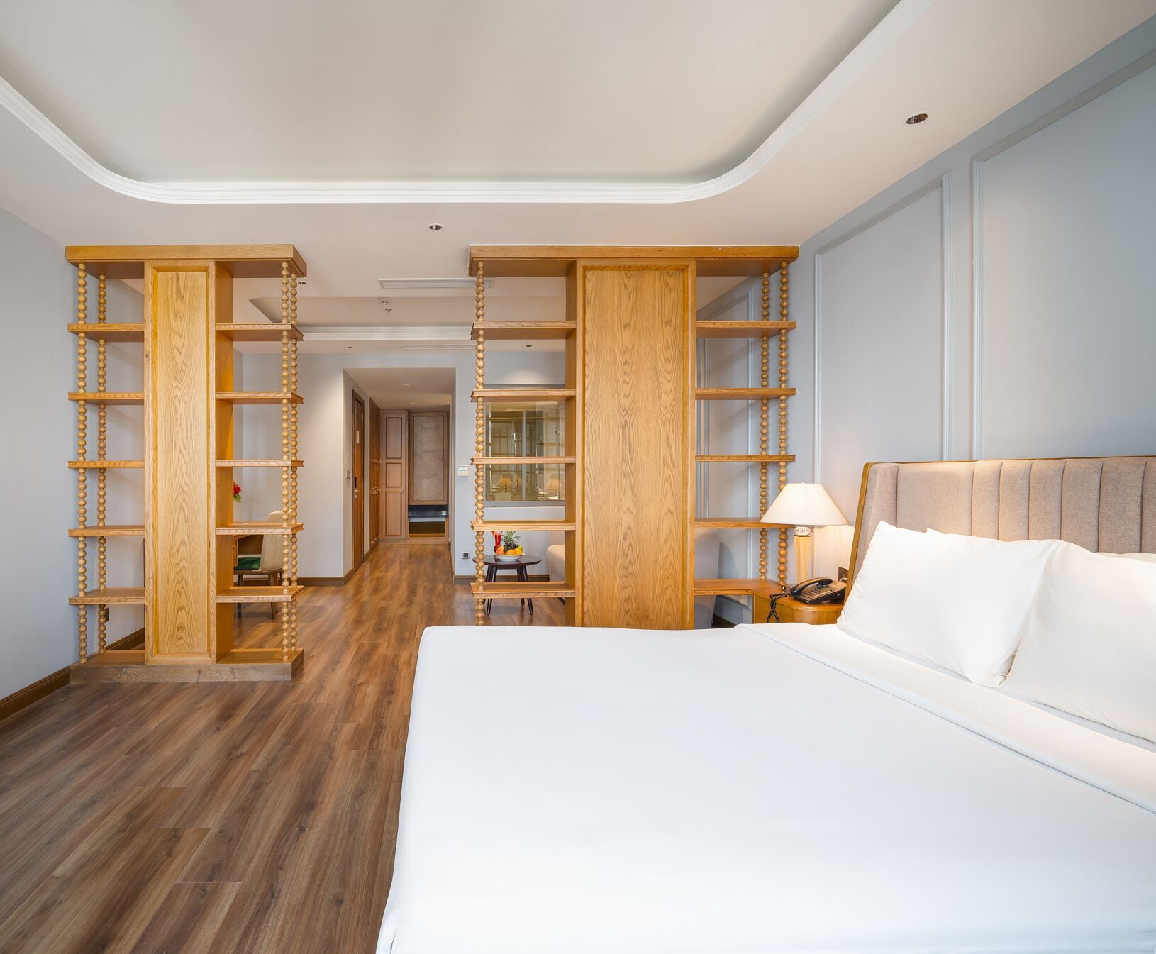A hotel room with a large bed, wooden shelving units, a small table with a lamp, and a view of the hallway.