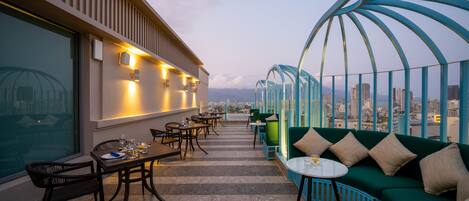 A rooftop terrace with a dining area, green seating, and a city view.