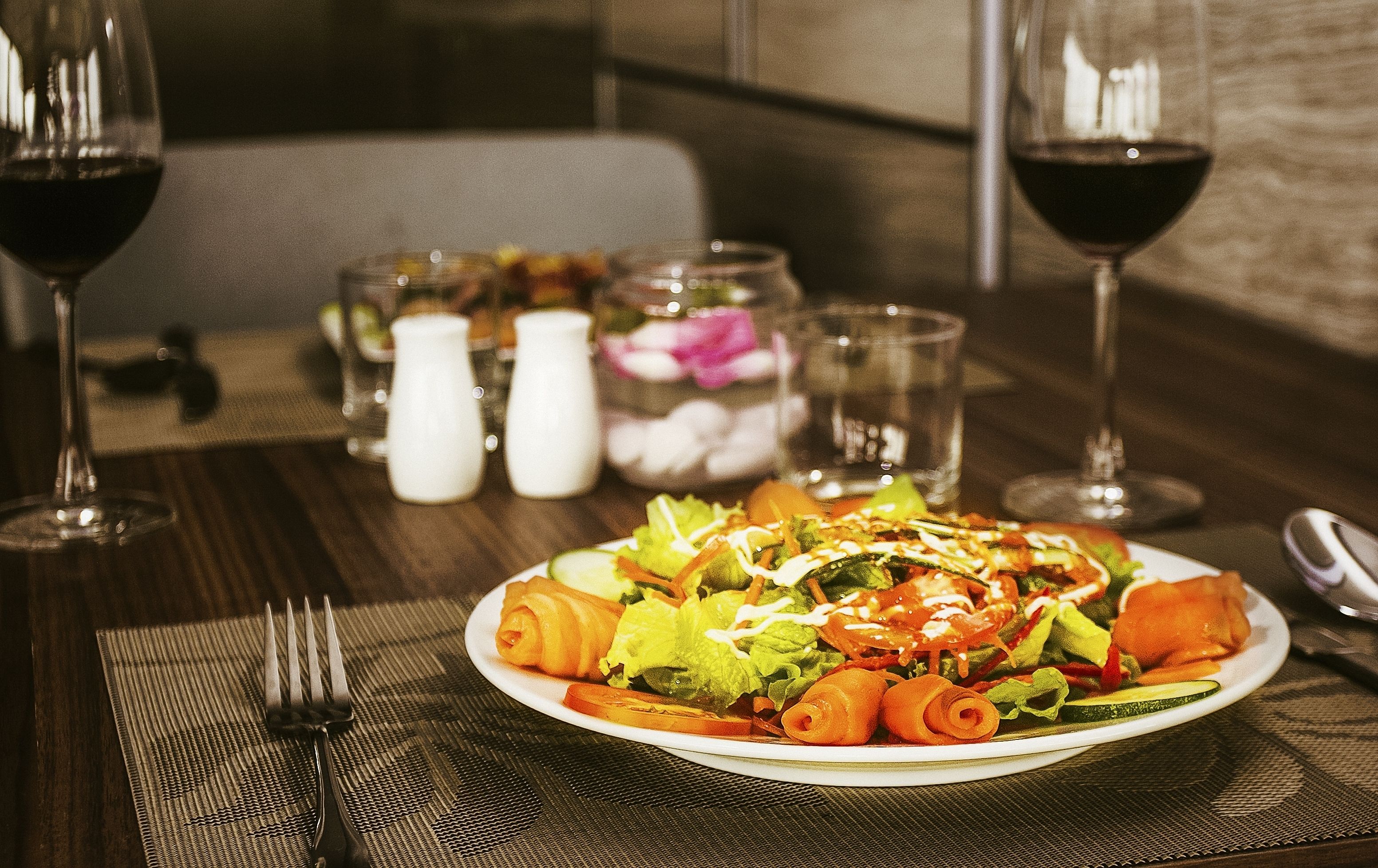 Una mesa de comedor con un plato de ensalada, dos copas de vino, saleros y pimenteros, y un florero con flores.