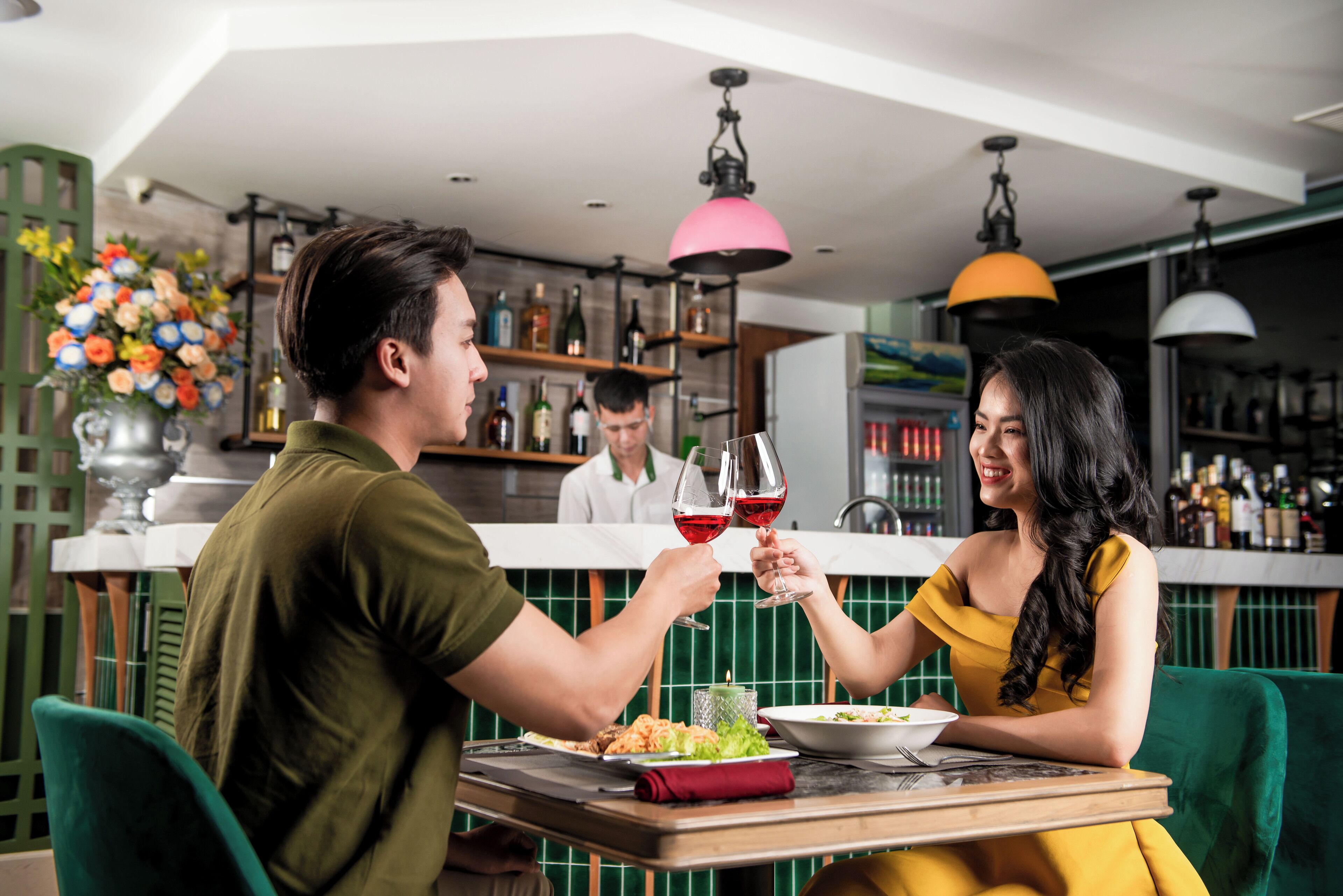 A dining area with a table set for two, a bar in the background, and a decorative vase of flowers.