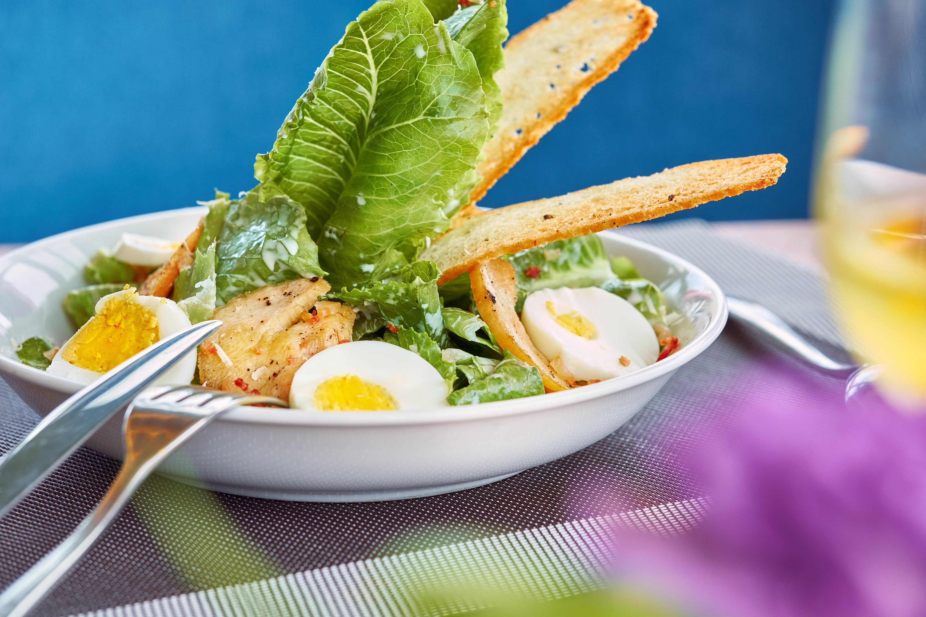 A bowl of salad with lettuce, eggs, and croutons on a table.