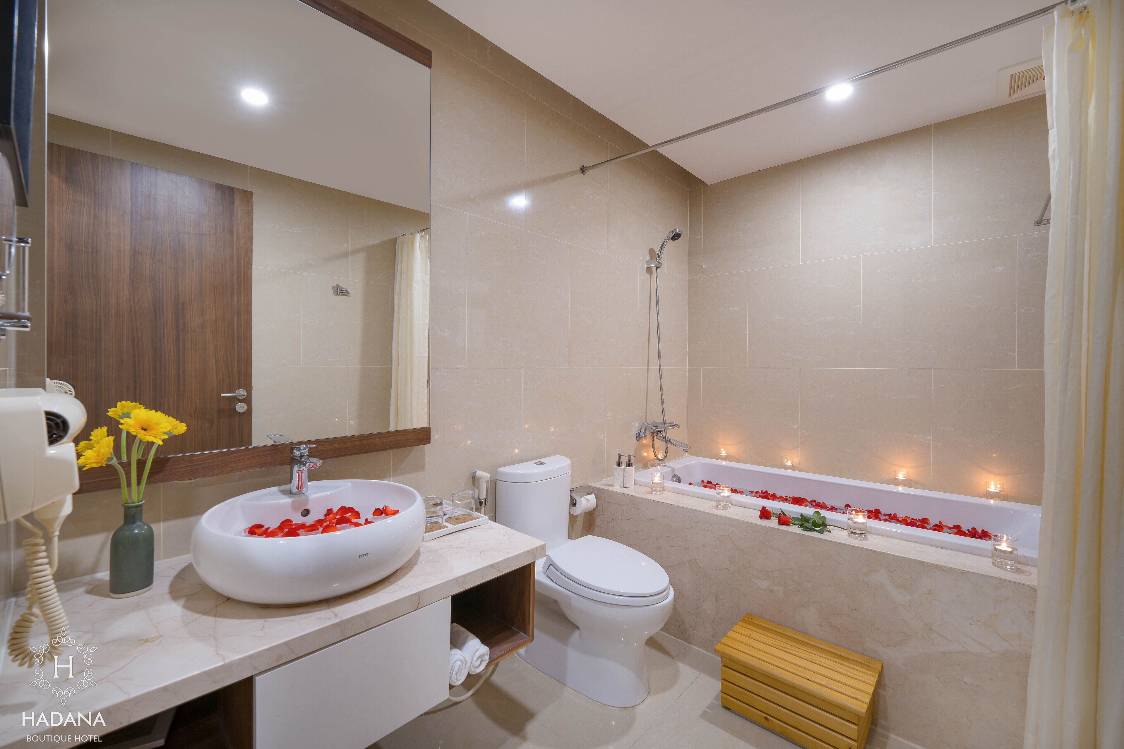 A modern bathroom with a white sink, toilet, and bathtub. There are flowers on the sink and candles in the bathtub.