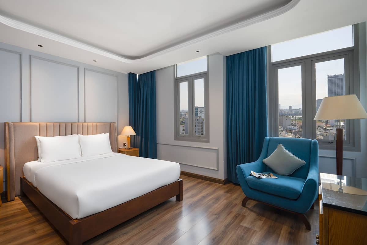 A hotel room with a large bed, a blue armchair, a nightstand with a lamp, and a city view through large windows.