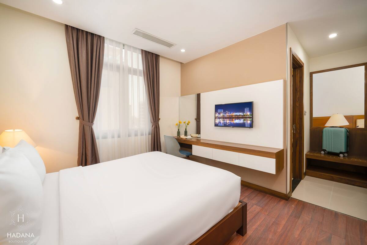 A hotel room with a bed, a desk with a TV, a mirror, and a suitcase.