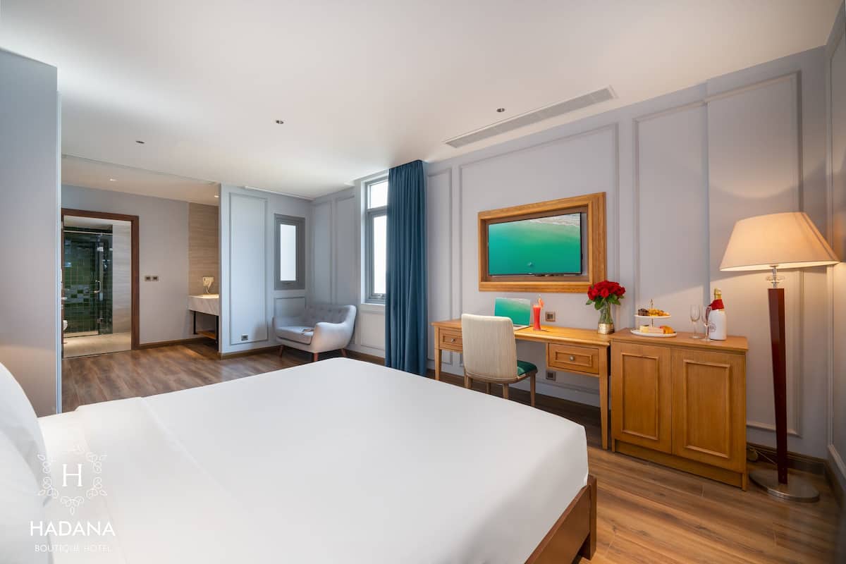A modern hotel room with a large bed, a desk, a chair, a vanity, and a television.