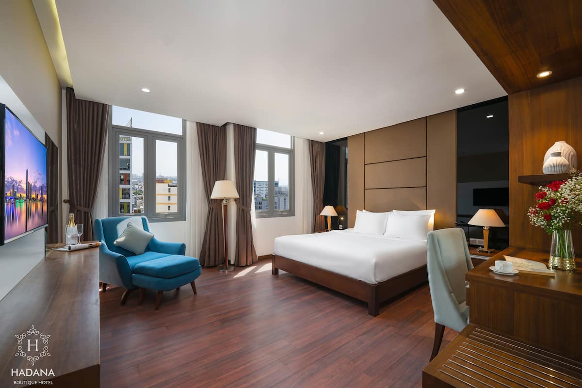 A modern hotel room with a large bed, a blue armchair, a desk, and a television.