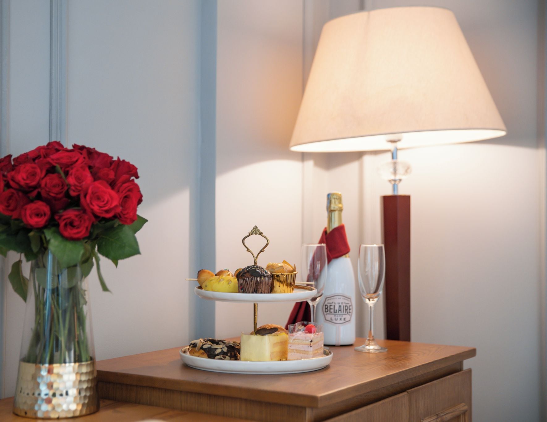 A table with a vase of red roses, a three-tiered dessert tray, a bottle of champagne, and a glass.