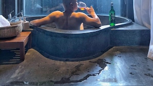 Una persona relajándose en una bañera con una botella de licor y una copa de vino, una ventana con vistas a bambú y árboles, y un taburete de madera rústica con una cesta tejida.