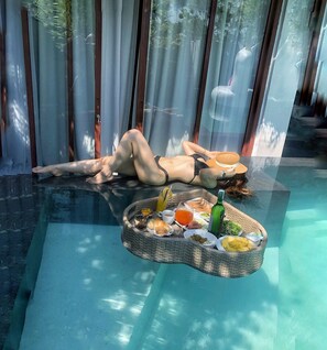 A person relaxing by a glass-bottomed pool with a wicker tray containing food and drinks. - Palm View Villa Hoi An (Da Nang)