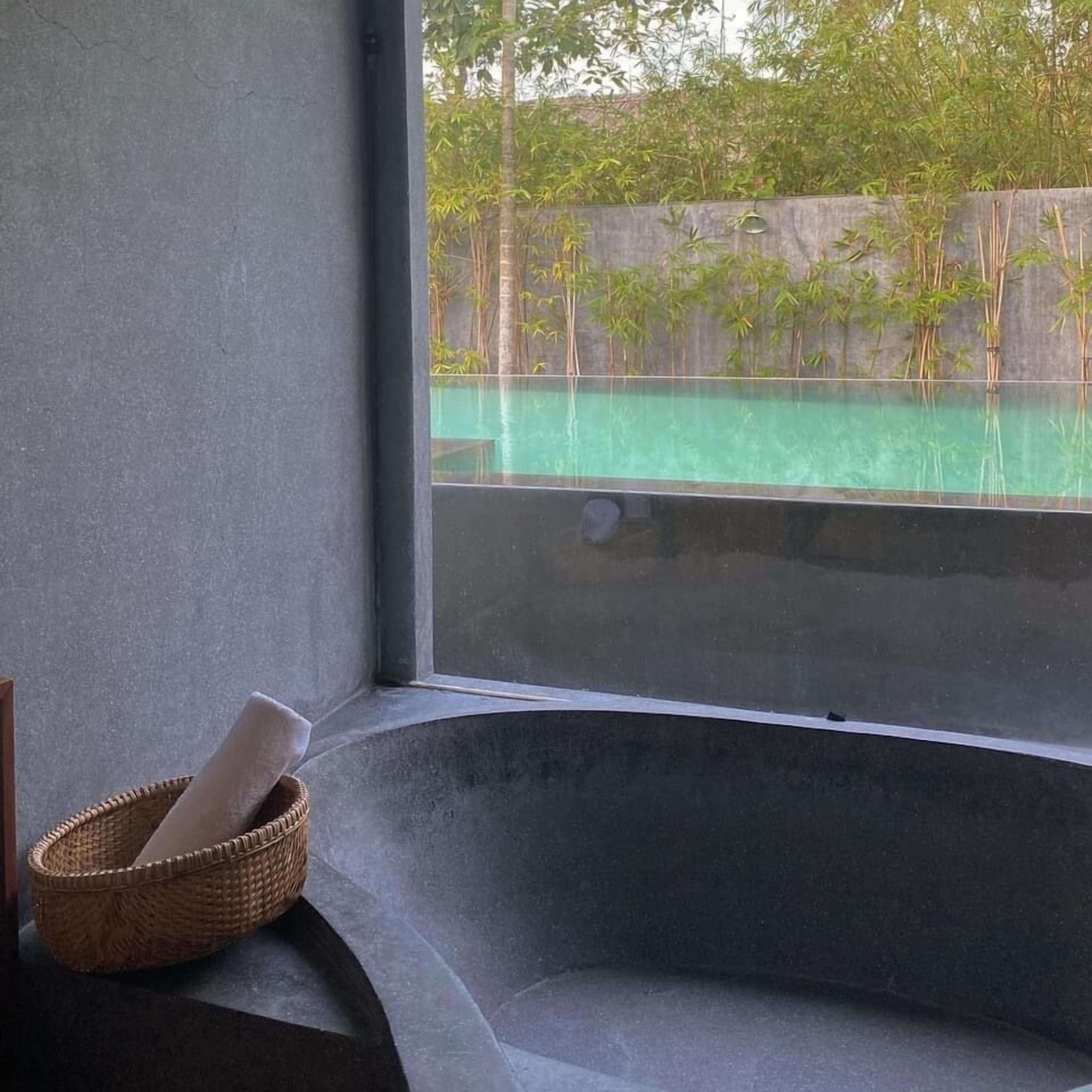 A corner of a room with a concrete bench, a wicker basket with a rolled-up mat, and a view of a bamboo fence and greenery outside.
