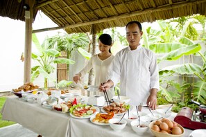 A dining table with various dishes and a food processor, surrounded by lush greenery.