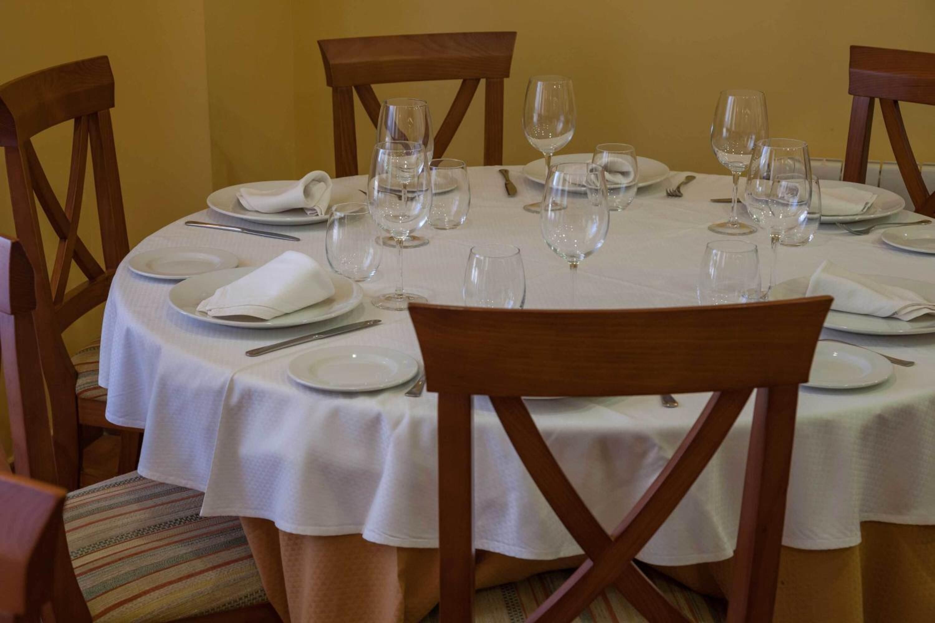 A round dining table set for six with white tablecloths, wooden chairs, and empty wine glasses.