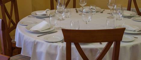 A round dining table set for six with white tablecloths, wooden chairs, and empty wine glasses.