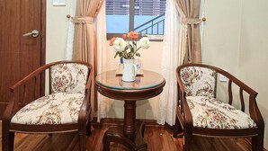 A room with two wooden chairs, a small round table with a vase of flowers, a standing fan, and a window with curtains. - Quan Quan Hotel (Da Nang)
