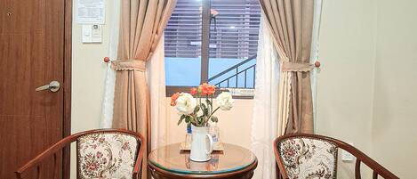 Una habitación con dos sillas de madera, una mesita redonda con un jarrón de flores, un ventilador de pie y una ventana con cortinas.