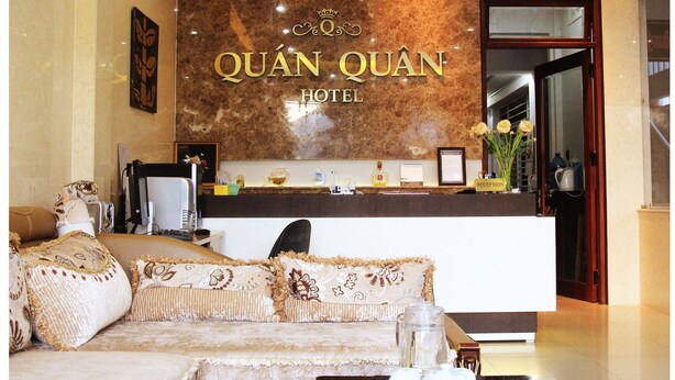 A hotel reception area with a large sign reading "QUÁN QUÁN HOTEL," a reception desk, a sofa with decorative pillows, and a coffee table with a bowl and flowers. - Quan Quan Hotel (Da Nang)