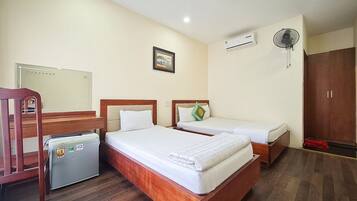 A hotel room with two beds, a small refrigerator, a wooden chair, a wardrobe, and a ceiling fan.