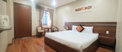A hotel room with a wooden floor, a bed with white bedding, a wooden headboard, a bedside table, a chair, a desk, a window with curtains, and a wall-mounted artwork.