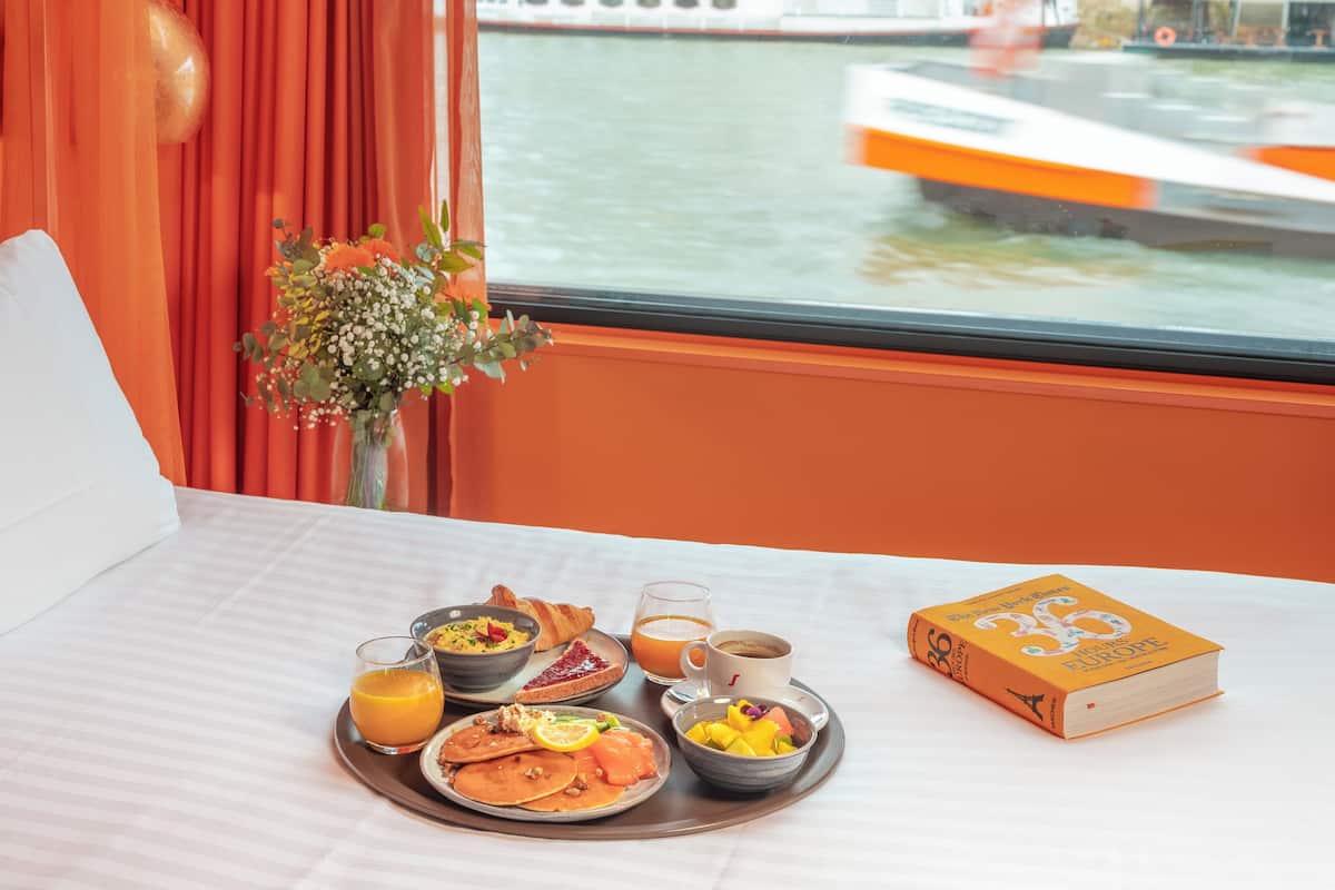 Ein Frühstückswagen mit Pfannkuchen, Obst und Kaffee, ein Buch mit dem Titel 'Europa' und Blick auf einen Fluss mit Booten.