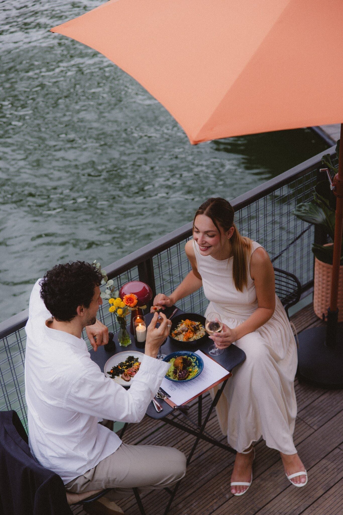 Un couple est assis à une table en plein air, devant un parasol, et profite de la vue sur l’eau.