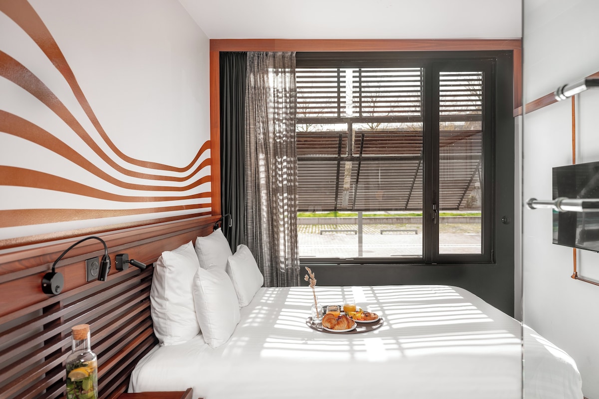 A hotel room with a bed, a bedside table with a bottle and a book, a TV on the wall, and a window with blinds.