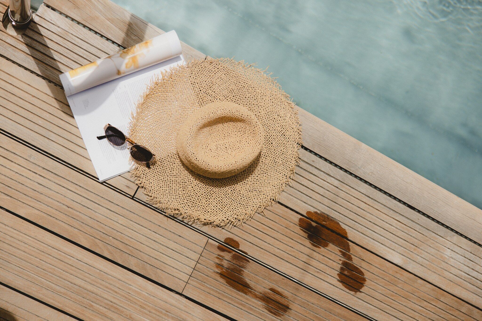 Un sombrero de paja, gafas de sol y una revista sobre una cubierta de madera junto a una piscina.