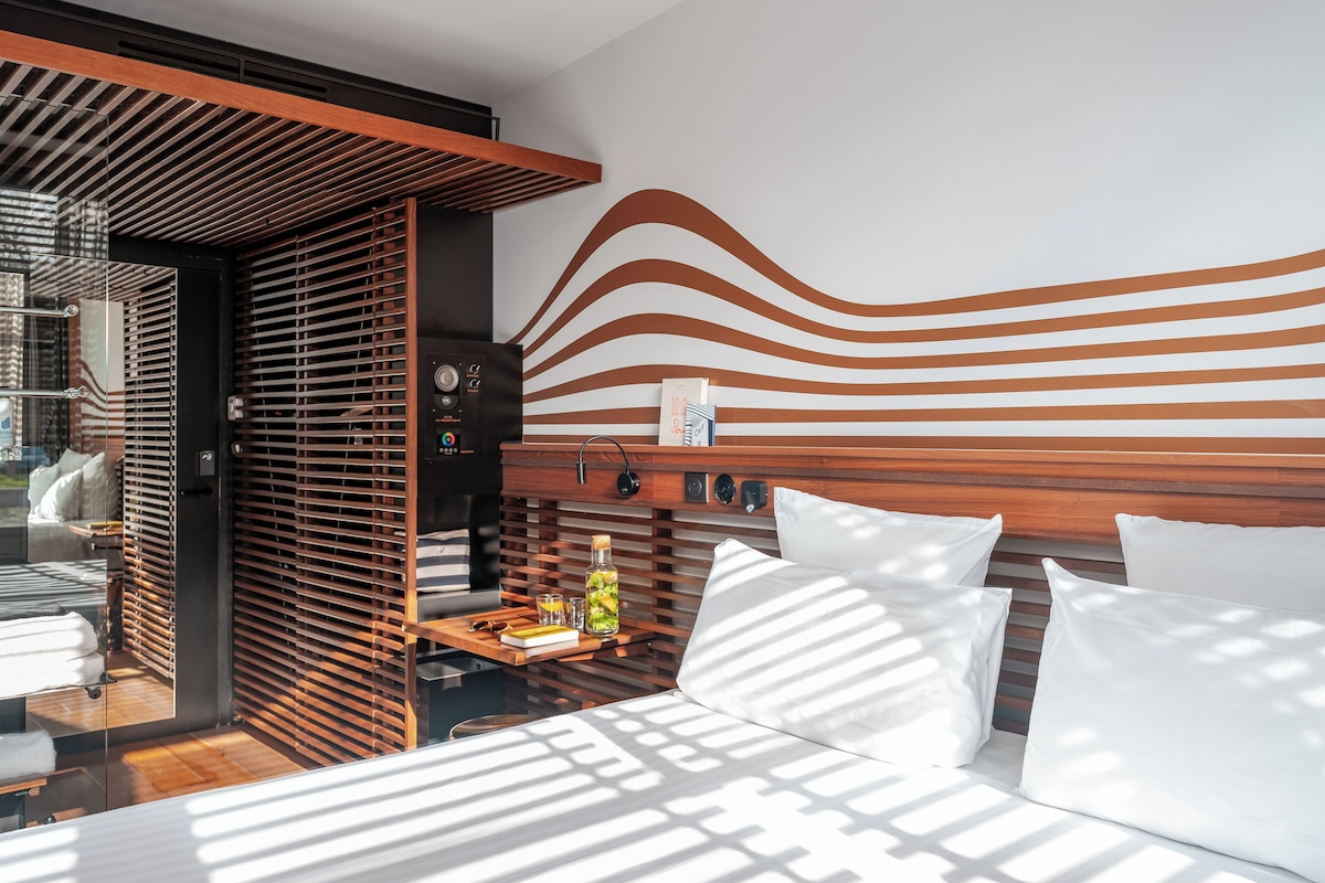 A modern hotel room with a bed, wooden headboard, and a wall with a wavy pattern. There is a small table with a bottle and glasses, and a window with blinds.