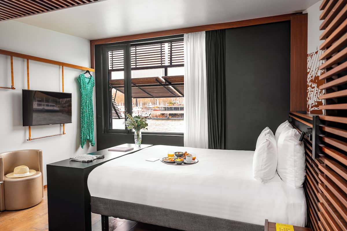 Ein modernes Hotelzimmer mit einem großen Bett, einem an der Wand befestigten Fernseher, einem kleinen Tisch mit Frühstücksangeboten und Blick auf Boote durch das Fenster.