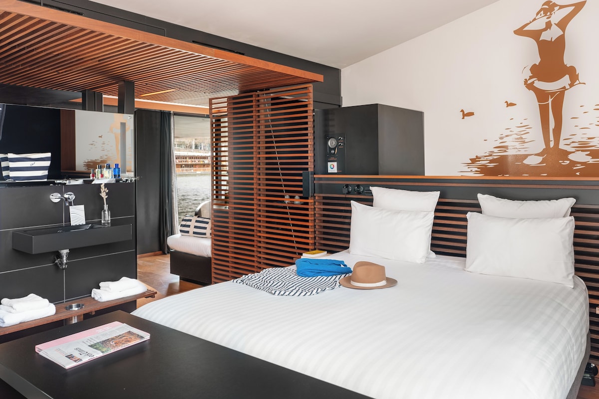 A modern hotel room with a large bed, a TV, a bathroom with a mirror, and a view of the water outside.