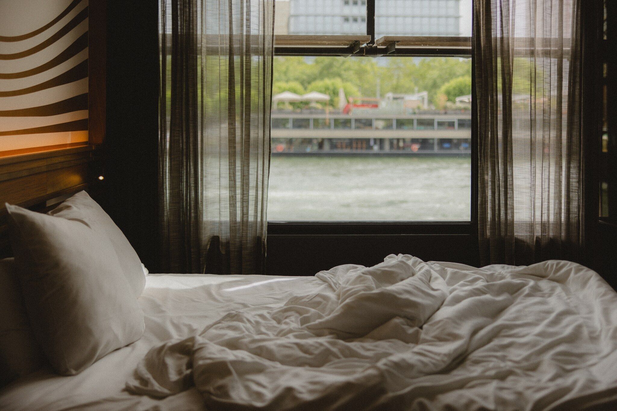 A bed with white bedding and a view of a river with a bridge and buildings in the background through sheer curtains.