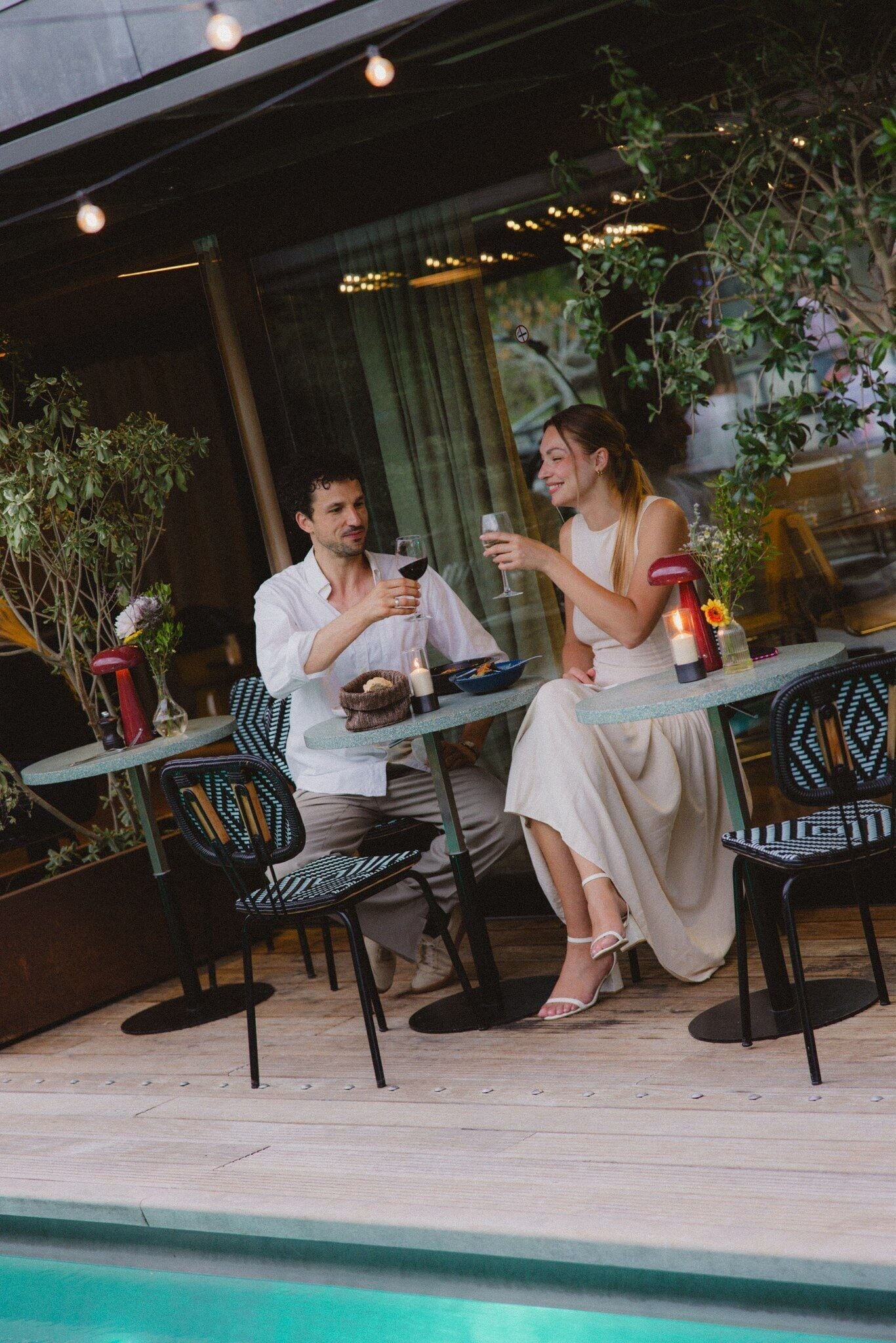 An outdoor dining area with two people seated at a table, a pool in the background, and potted plants around.
