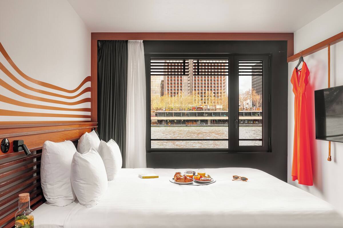 Ein Hotelzimmer mit Bett, Fenster mit Jalousien, Fernseher und Frühstückswagen auf dem Bett.