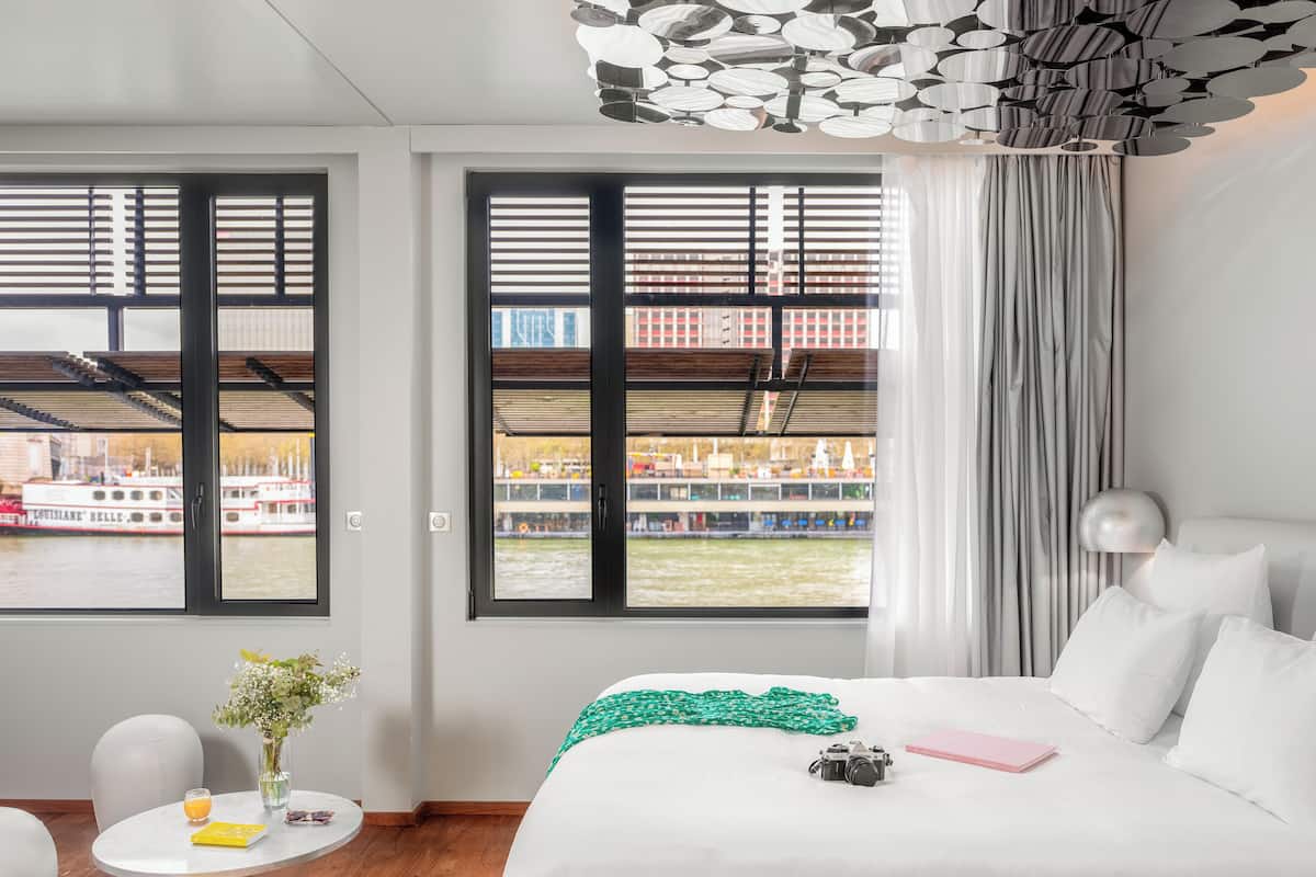 Ein Hotelzimmer mit einem großen Bett, einem kleinen runden Tisch mit Blumen in einer Vase, einer Kamera und einem rosafarbenen Buch.