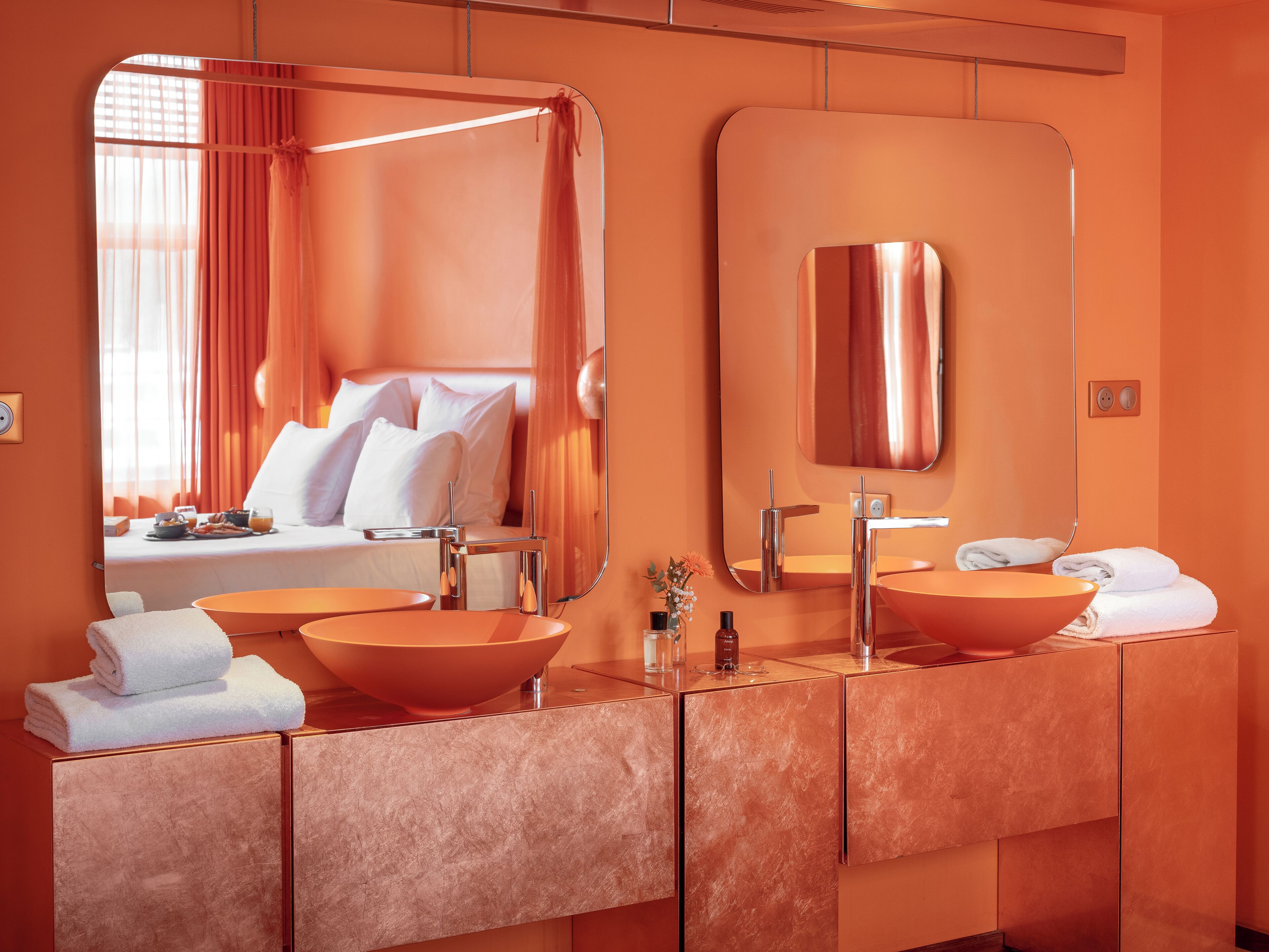 Ein modernes Badezimmer mit einem großen Spiegel, zwei Waschbecken und einem durch das Fenster gut erkennbaren Bett.